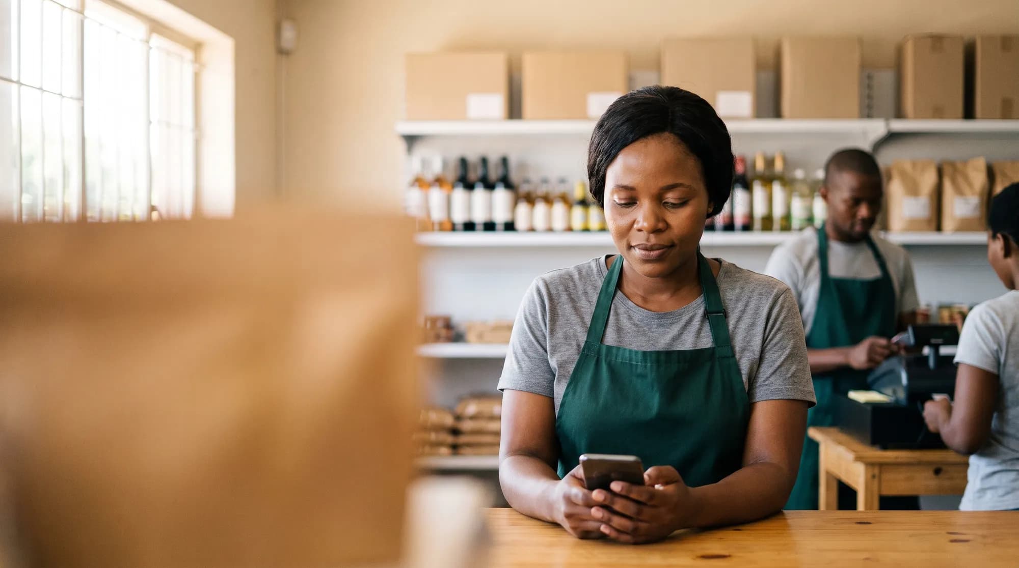 A grocery store owner checks stock on her phone at the shop counter while a colleague serves a customer behind her.